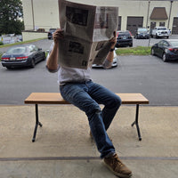 Garden bench by Treeboard with white oak seat and cast-iron legs. On the seat is a man reading The New York Times newspaper.