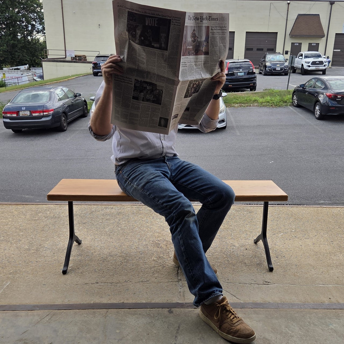 Garden bench by Treeboard with white oak seat and cast-iron legs. On the seat is a man reading The New York Times newspaper.
