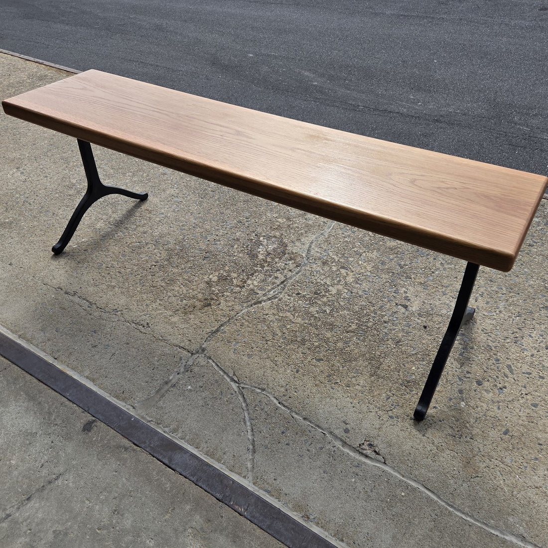 Garden bench by Treeboard with white oak seat and cast-iron legs. Below is concrete, with asphalt in the background.