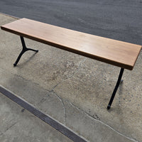 Garden bench by Treeboard with white oak seat and cast-iron legs. Below is concrete, with asphalt in the background.