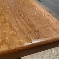Garden bench by Treeboard with white oak seat and cast-iron legs. Rain has fallen onto the benchtop, but the polyurethane finish is repelling it, causing the water to bead up.